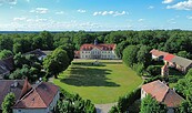 Luftaufnahme vom Schloss Grochwitz mit rotem Dach, großer Wiese, Nebengebäuden und dichtem Wald unter blauem Himmel., Foto: Bettina Unterkofler, Lizenz: Bettina Unterkofler
