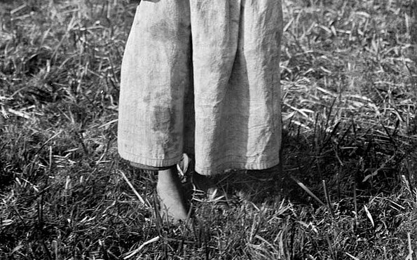 Ein Schulmädchen in wendischer Tracht steht mit Schulbüchern in der Hand barfuß auf einer Wiese. Altes schwarz-weiß Foto, Foto: Denis Galloway, Lizenz: Ethnographical Museum of Transylvania, Romania