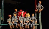 Sieben sitzende Puppen mit blauen Overalls und braunen Schuhen vor einem roten Kaffeekannen-Requisit auf einer Bühne, Foto: Johann Karl, Lizenz: DAS WEITE THEATER