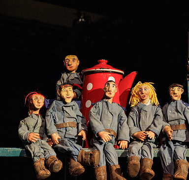 Sieben sitzende Puppen mit blauen Overalls und braunen Schuhen vor einem roten Kaffeekannen-Requisit auf einer Bühne, Foto: Johann Karl, Lizenz: DAS WEITE THEATER