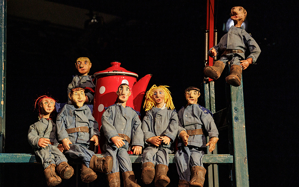 Sieben sitzende Puppen mit blauen Overalls und braunen Schuhen vor einem roten Kaffeekannen-Requisit auf einer Bühne, Foto: Johann Karl, Lizenz: DAS WEITE THEATER