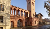 Rötlich verputzte dreibogige Toreinfahrt mit Arkadengang und Turm neben hellem Gebäude vor blauem Himmel., Foto: Sophie Soike, Lizenz: PMSG