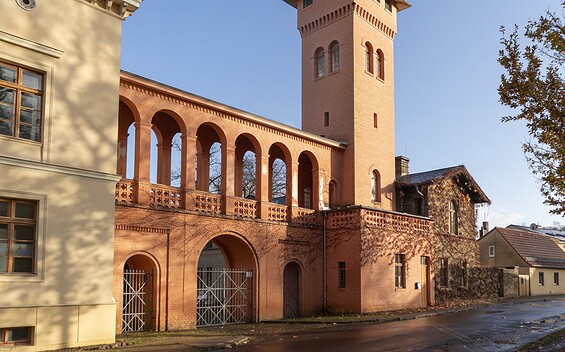 Rötlich verputzte dreibogige Toreinfahrt mit Arkadengang und Turm neben hellem Gebäude vor blauem Himmel., Foto: Sophie Soike, Lizenz: PMSG
