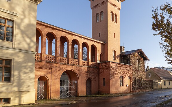 Rötlich verputzte dreibogige Toreinfahrt mit Arkadengang und Turm neben hellem Gebäude vor blauem Himmel., Foto: Sophie Soike, Lizenz: PMSG