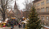 Menschen auf einem Weihnachtsmarkt zwischen Backsteingebäuden, kahlen Bäumen und beleuchteten Tannen; im Hintergrund ein Kirchturm., Foto: André Stiebitz, Lizenz: PMSG