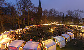 Beleuchteter Weihnachtsmarkt am Abend vor einer Kirche, mit vielen Besuchern, weißen Zelten und kahlen Bäumen., Foto: André Stiebitz, Lizenz: PMSG