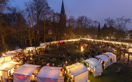 Beleuchteter Weihnachtsmarkt am Abend vor einer Kirche, mit vielen Besuchern, weißen Zelten und kahlen Bäumen., Foto: André Stiebitz, Lizenz: PMSG