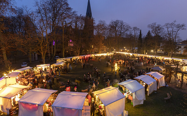 Beleuchteter Weihnachtsmarkt am Abend vor einer Kirche, mit vielen Besuchern, weißen Zelten und kahlen Bäumen., Foto: André Stiebitz, Lizenz: PMSG