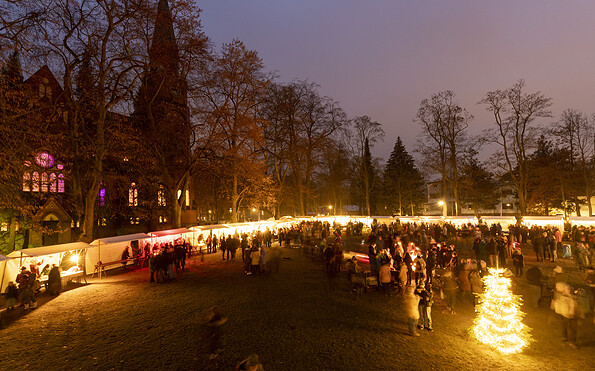 Abendlicher Weihnachtsmarkt mit beleuchteten Ständen und vielen Menschen vor einer Kirche zwischen kahlen Bäumen., Foto: André Stiebitz, Lizenz: PMSG