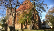 Backsteinkirche mit Turm und rotem Ziegeldach, umgeben von herbstlichen Bäumen; im Vordergrund eine Steinskulptur., Foto: Anja Warning, Lizenz: Anja Warning