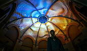 Blau-gelb beleuchtetes, gewölbtes Glasdach in einem Innenhof; unten rechts steht eine Person., Foto: Moritz Richartz
