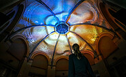 Blau-gelb beleuchtetes, gewölbtes Glasdach in einem Innenhof; unten rechts steht eine Person., Foto: Moritz Richartz