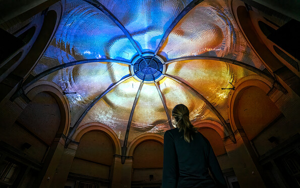 Blau-gelb beleuchtetes, gewölbtes Glasdach in einem Innenhof; unten rechts steht eine Person., Foto: Moritz Richartz