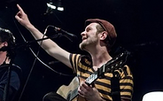 Mann mit gestreiftem T-Shirt und Mütze spielt Gitarre und zeigt mit ausgestrecktem Arm nach oben auf einer dunklen Bühne, Foto: Peter Wolter, Lizenz: Peter Wolter
